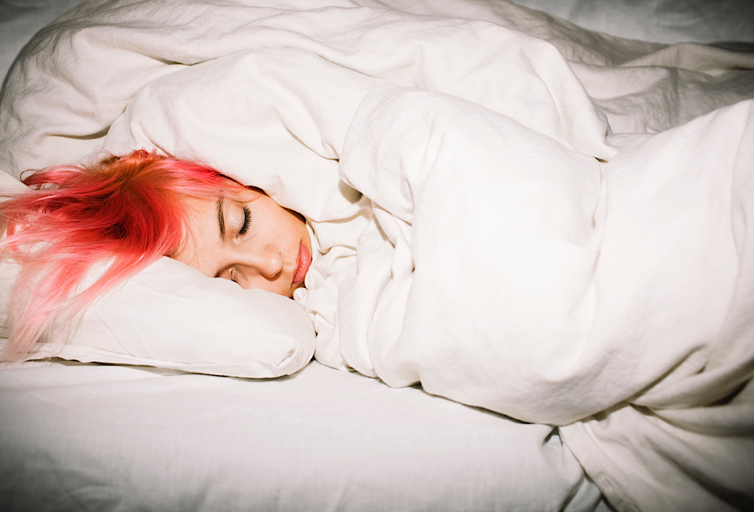 Young woman with pink hair sleeping in bed wrapped up in a white comforter and sheets.