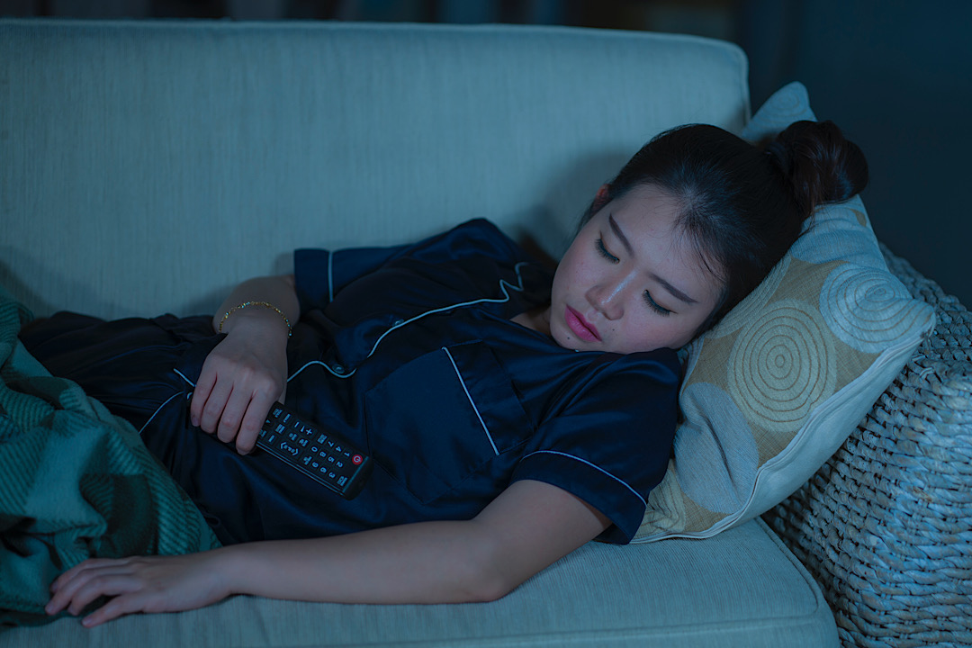 Young Asian woman asleep on couch with TV on.