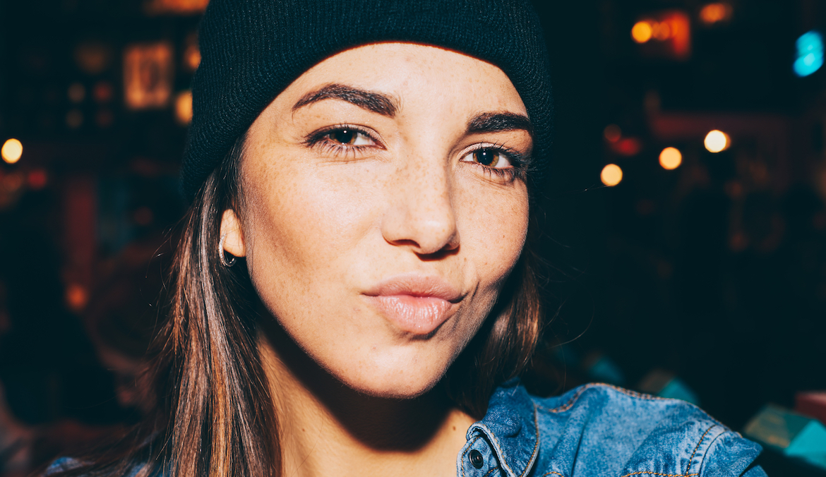 Photo of lovely brunette with plump lips and healthy skin wearing denim jacket and black beanie hat looking at camera, lights on dark background.