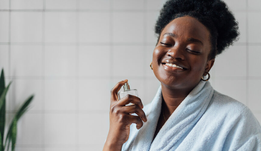 A young smiling woman sprays non-toxic perfume onto her neck while wearing a bath robe inside of a white bathroom.