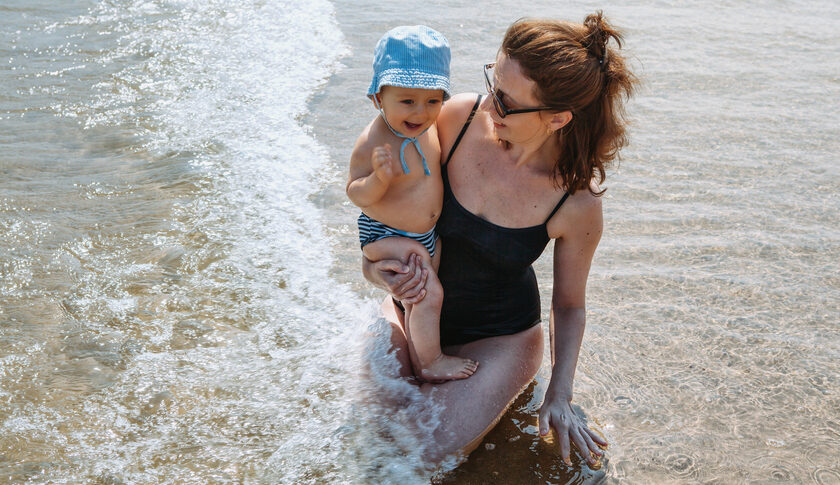 A new mom wearing a postpartum bathing suit holds her newborn baby while playing in the ocean near the shore.