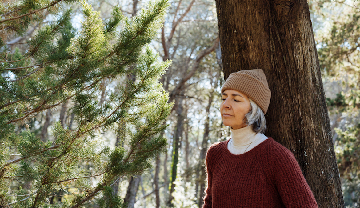 woman in sweater and hat closing eyes and leaning on tree trunk while resting and connecting with nature in forest