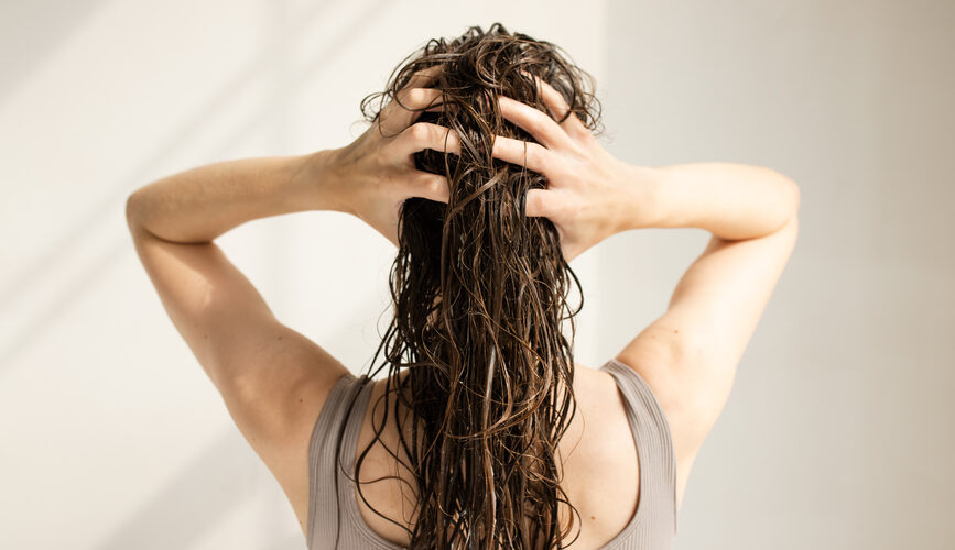 A woman runs her fingers through her long, wet hair while standing in front of a plain wall.