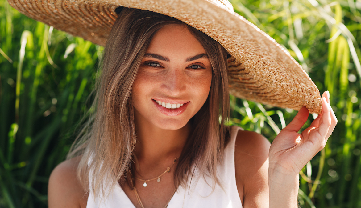 Beautiful blond woman in straw hat looking at camera and smiling while sitting outdoors