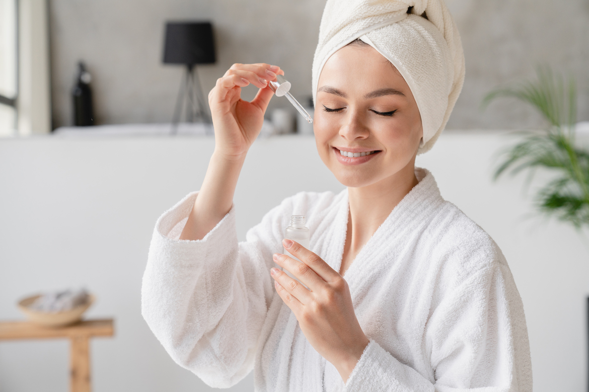 Young white woman in bath spa towel applying rejuvenation moisturizing serum on her face with her eyes closed at home after hot bath and shower. Beauty treatment concept