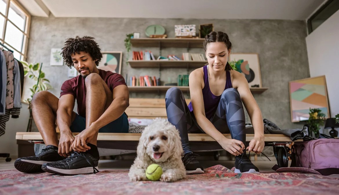 Young multi-ethnic couple sitting on bed at apartment and preparing for jogging, fluffy dog sitting near them