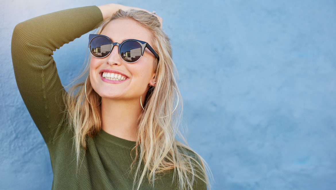 Close up shot of stylish young woman in sunglasses smiling against blue background. Beautiful female model with copy space.