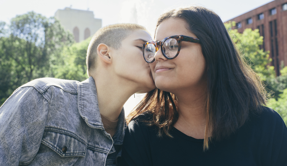 A young lesbian couple outdoors, one woman kisses the other on the cheek