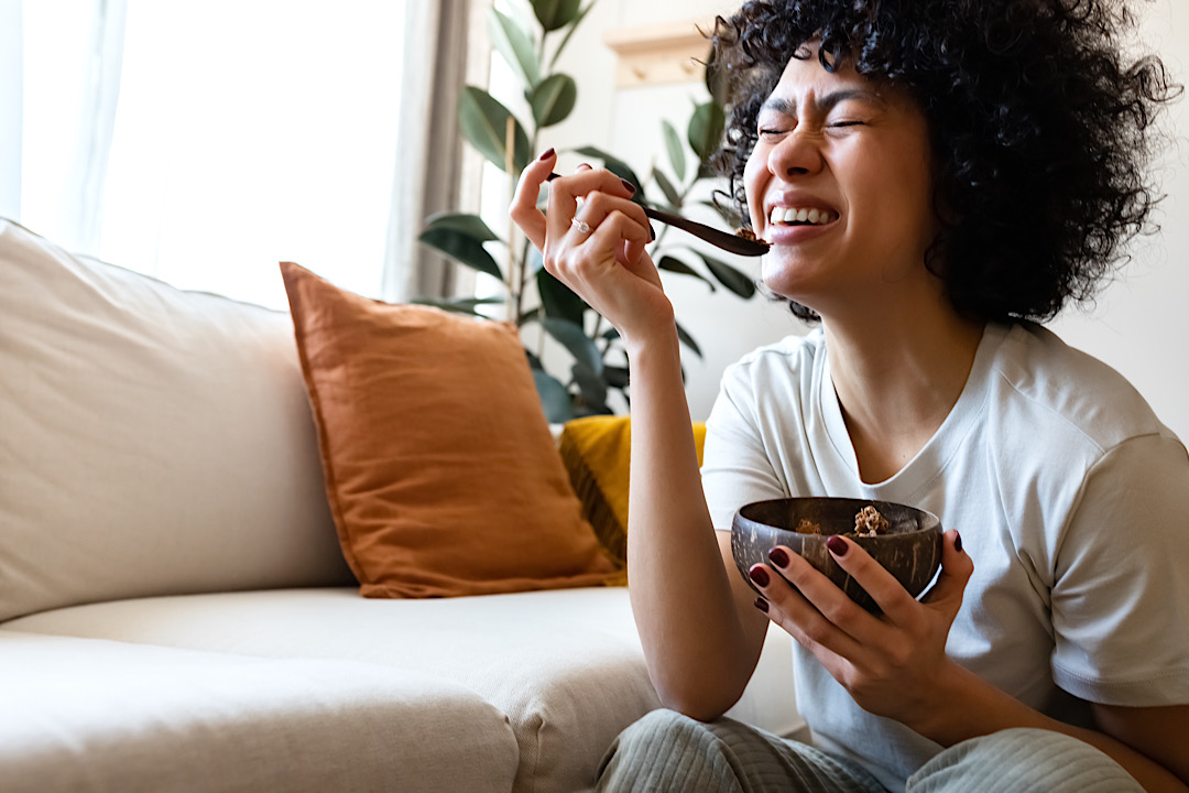 Young Black woman laughing and having fun while eating breakfast at home in her living room.