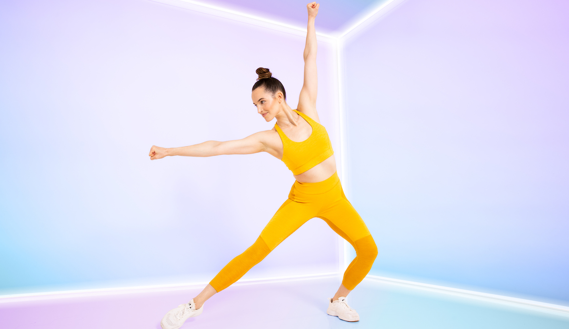 A woman in a pastel lit room wearing an orange sherbert colored exercise outfit with her arms and legs out in a strong fitness stance.