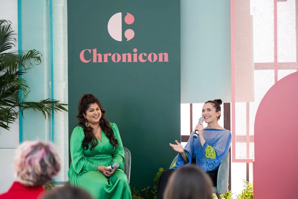 Nitika Chopra and Alyson Stoner sit in front of a Chronicon panel background, speaking to the audience. 