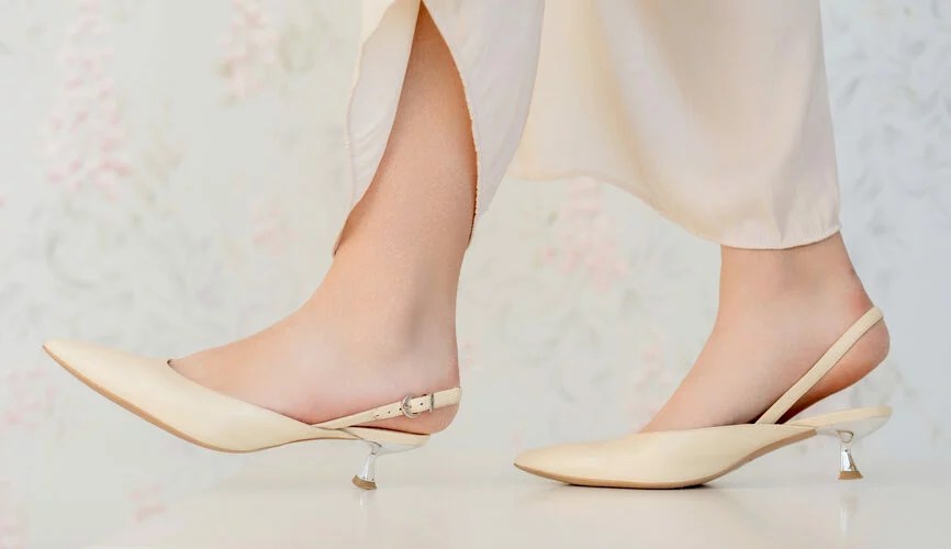 Women's feet wearing a pair of light-colored slingback shoes in front of a floral wallpaper background.