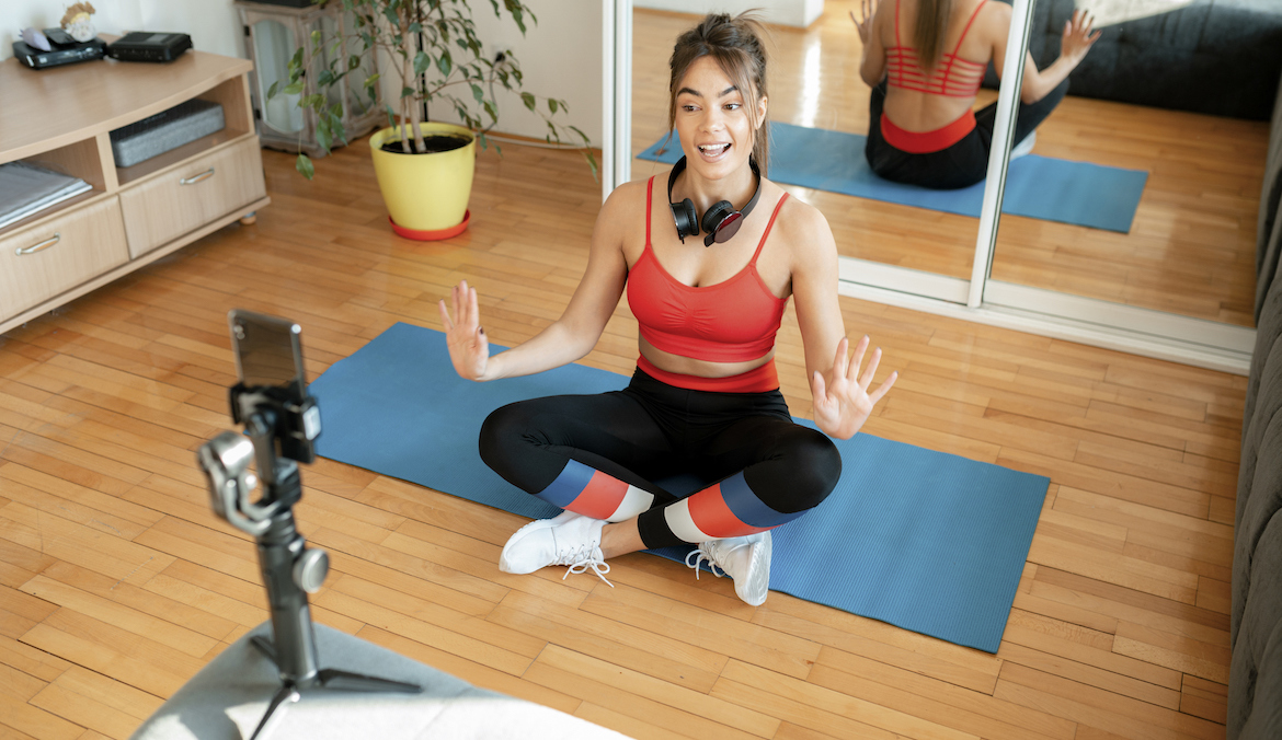 A woman in athletic gear sitting on a yoga mat talking into a smartphone camera propped up on a tripod.