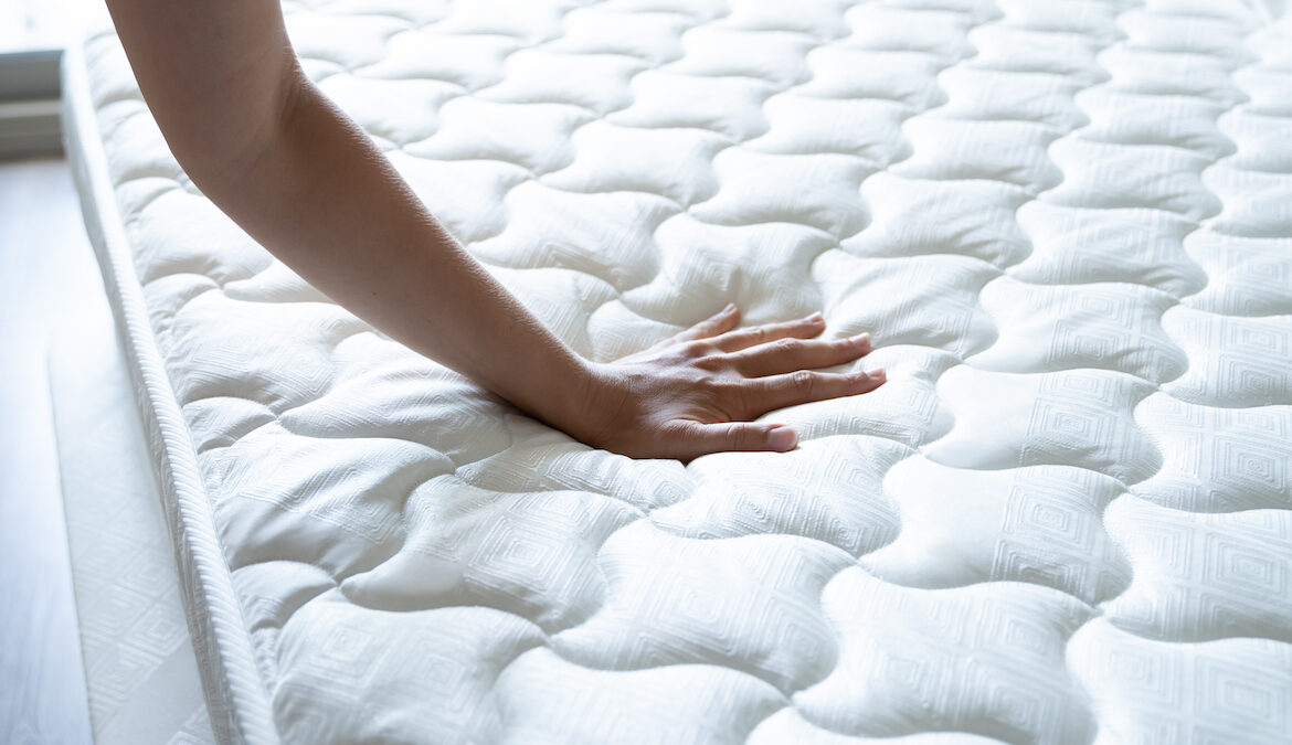 A hand presses down into a plush quilted mattress topper in a well-lit bedroom.