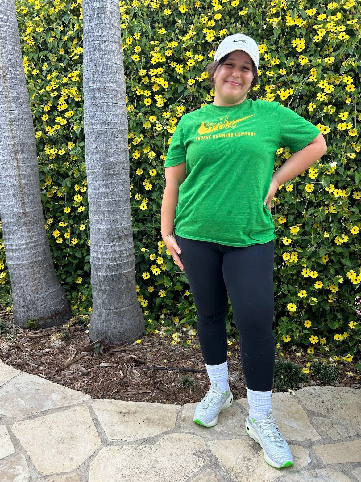 A woman wearing a white hat, green t-shirt with a yellow Nike swoosh logo, black leggings, white tube socks, and gray, white, and green tennis shoes stands in front of a green vine with yellow flowers.