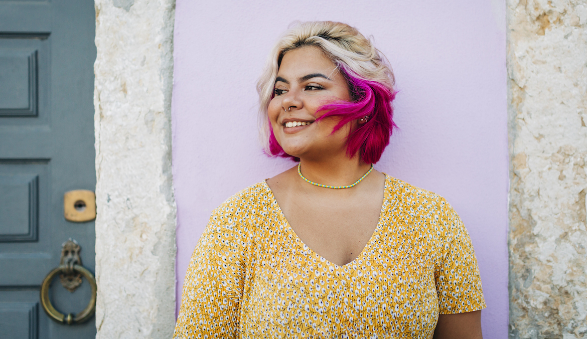 A blonde woman with pink hair stands and smiles in front of a house in Portugal.