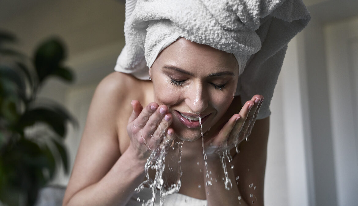 Woman splashes her face with ice water while wearing a bath towel on her head, standing in a bathroom.