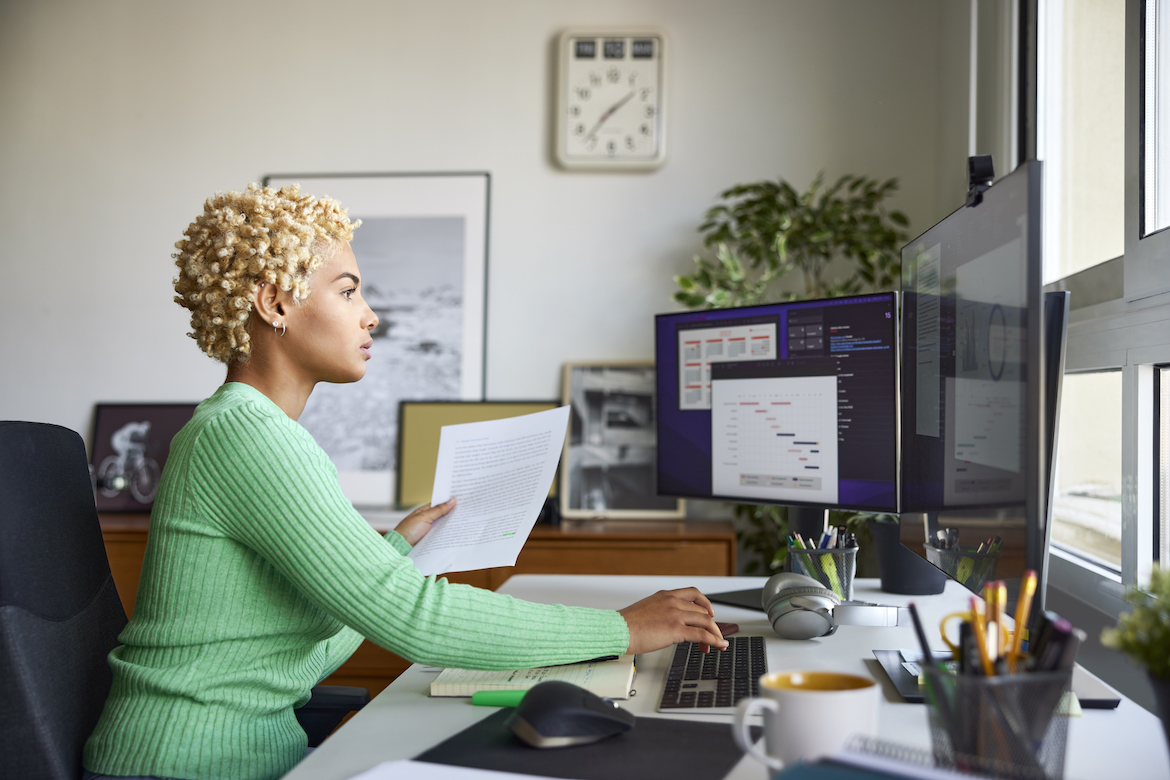 Side view of confident businesswoman holding document while using computer. Blond Afro female entrepreneur is working at home office. She is sitting at desk in apartment.