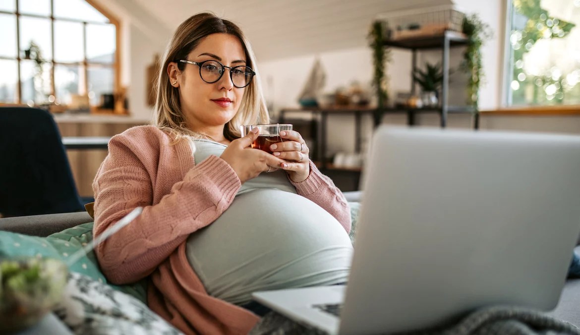 Young pregnant woman drinking raspberry leaf tea at home, sitting in sofa while having a video conference, using laptop.
