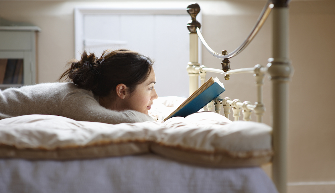 A woman lies near the foot of a bed and reads a book.