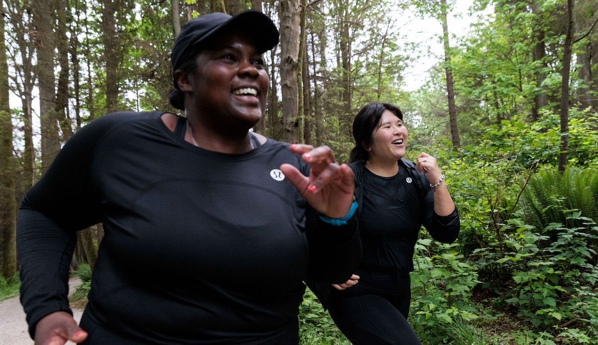 Two larger bodied women smiling while running through a lush green forest.