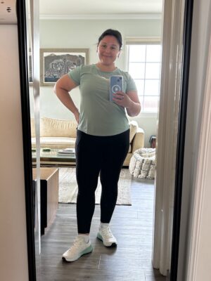 A woman in black leggings and a green shirt taking a selfie in a mirror, showing off a pair of white running shoes.