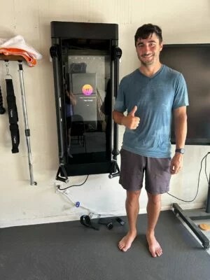 A sweaty man standing next to a fitness machine giving a thumbs up.