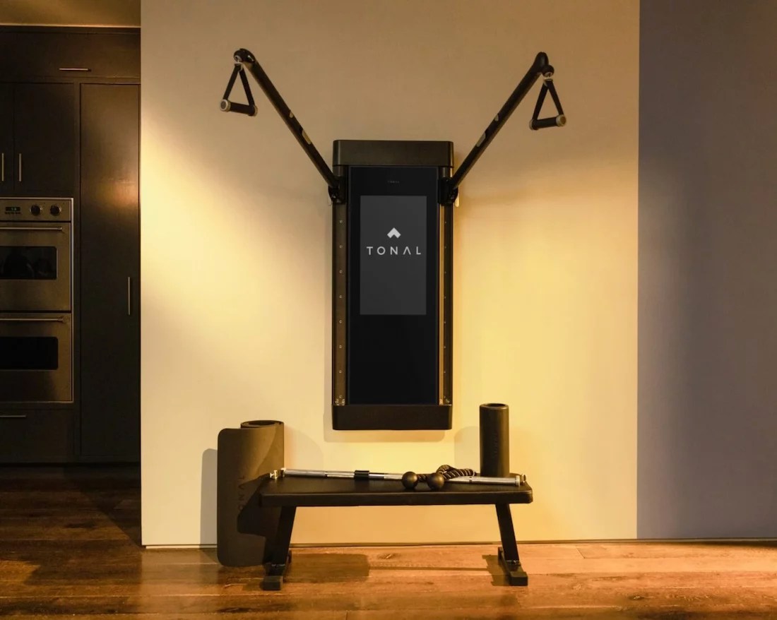 The Tonal home gym, showing the machine on the wall with extended arms, a bench, a foam roller, a rope, and a barbell.