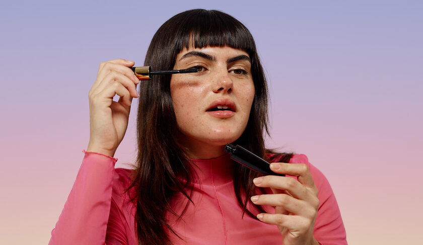 A woman with bangs applies mascara to her eyelashes.