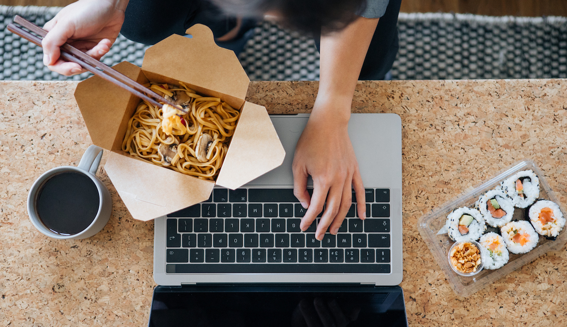 Top view of young woman eating takeaway Asian food while touching keyboard on the laptop, which she's going to need to clean
