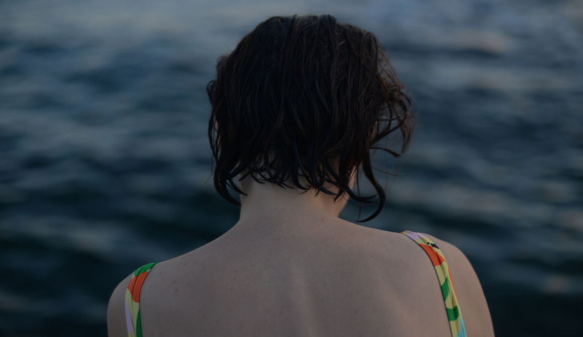 The back of a woman wearing a bathing suit looking out over the ocean, with a darker, ominous tinge over the image.