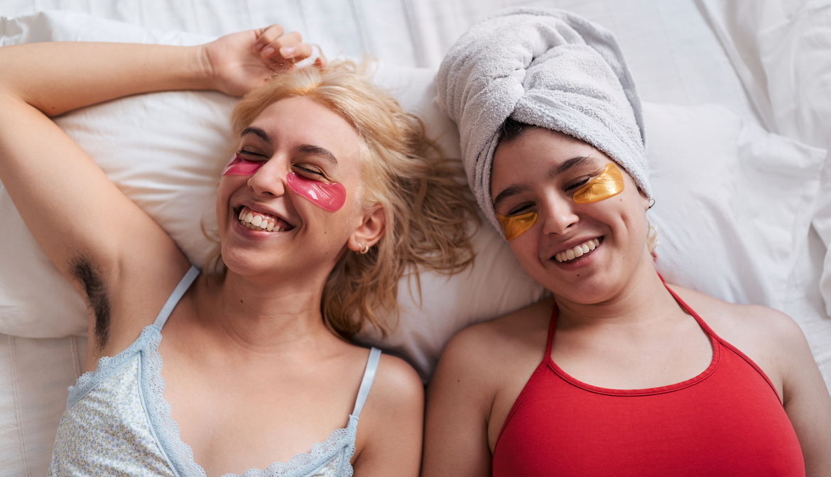 Two young natural women lying in bed laughing with colored beauty patches under their eyes.