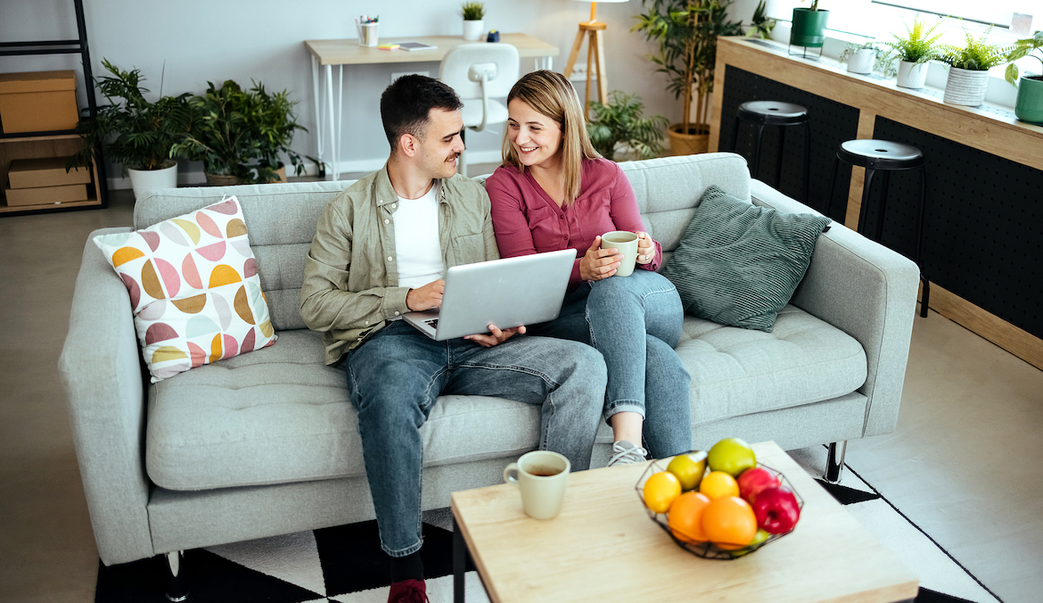 Couple sitting on a sofa, using a laptop, and the woman is drinking coffee