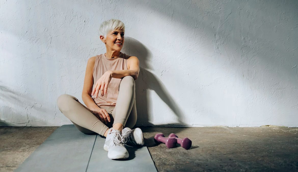A senior woman with cropped gray hair sitting against a wall on a yoga mat with hand weights beside her, to illustrate exercise advice for seniors