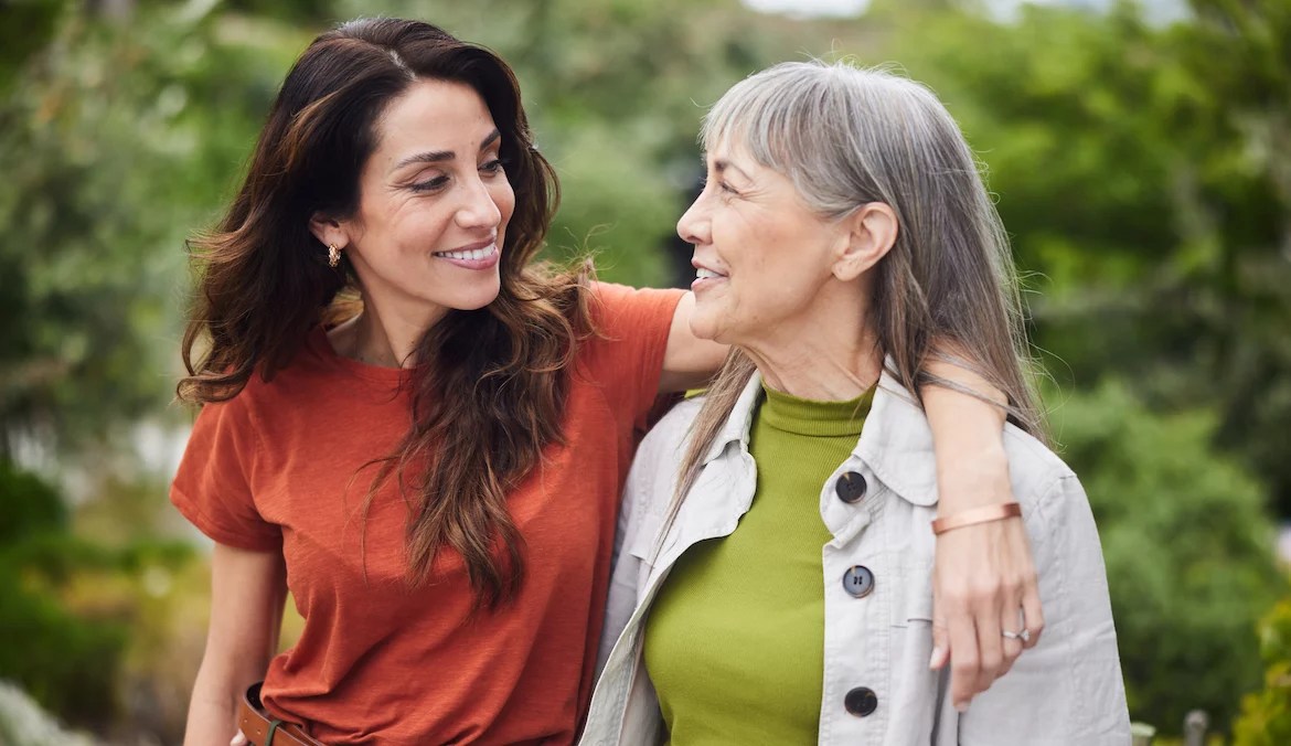 Woman standing arm in arm with her senior mother and smiling while standing together outside in a yard at home