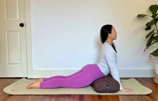 a female yoga teacher with long dark hair shows how to do resting cobra, a restorative version of the cobra pose in yoga, using a bolster under her hips