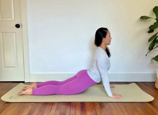 woman yoga teacher with long dark hair wearing bright purple leggings demonstrates how to do the cobra pose in yoga