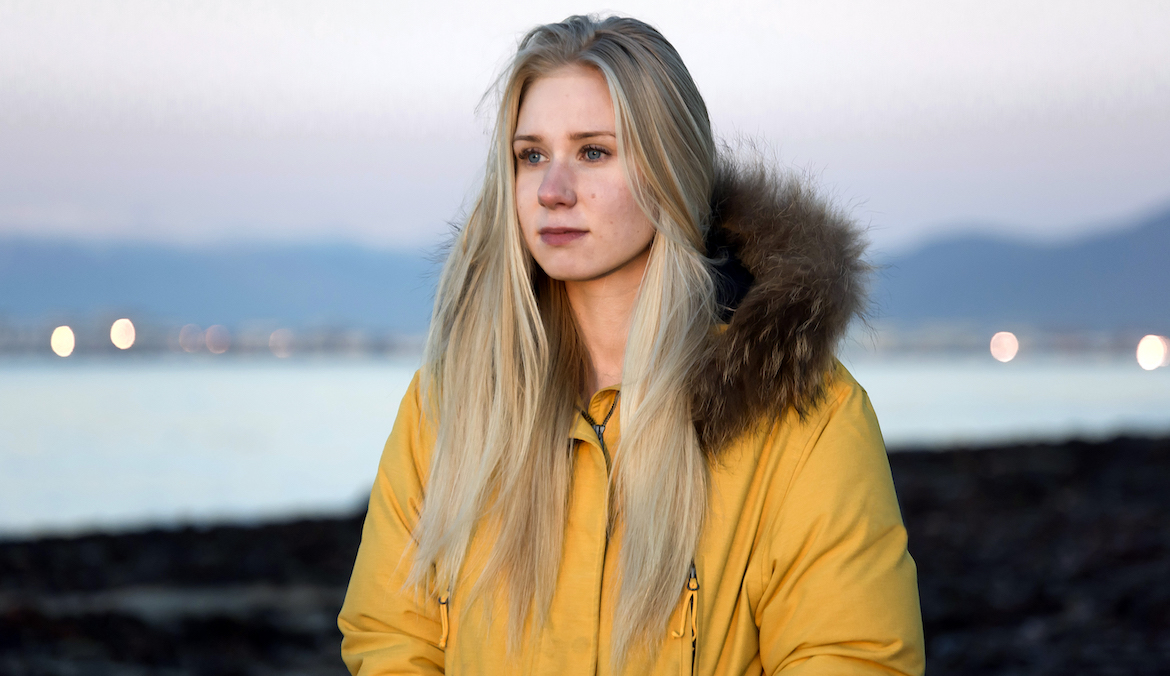 Portrait of a beautiful Icelandic blonde woman, wearing a yellow outdoor winter puffer jacket, walking along the beach at sunset. Belt of Venus visible on the horizon.