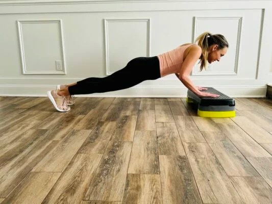female PT with a blond ponytail wearing a tank top and leggings shows how to do correct incline push-ups
