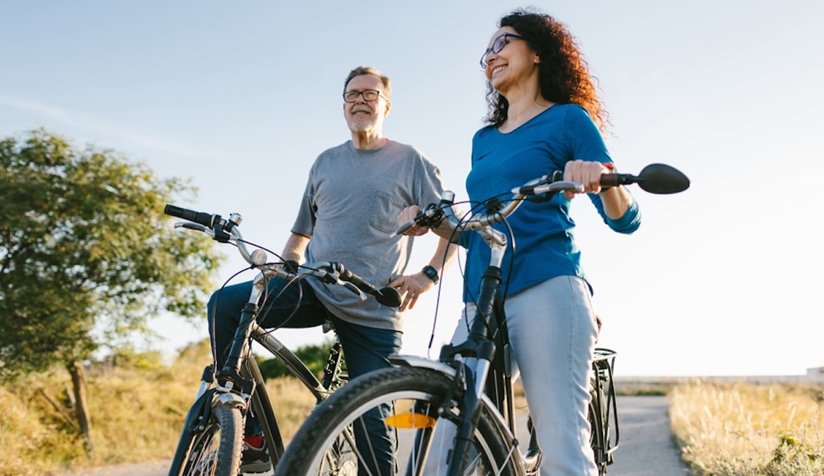 A middle-aged couple is riding a bicycle in the countryside. It is a sunny day in Spring at the end of the afternoon, at sunset. They are both smiling and having a good time together.