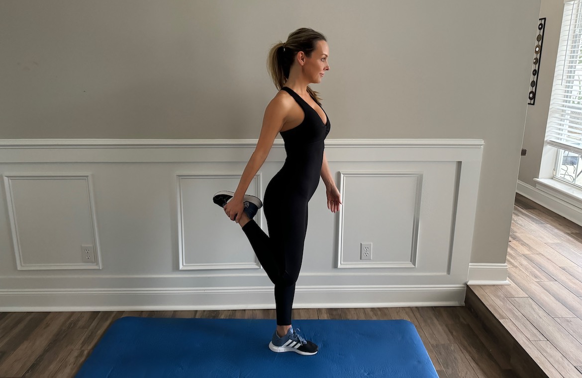 female PT with a long blond ponytail wearing black workout onesie shows the correct form for the standing quad stretch