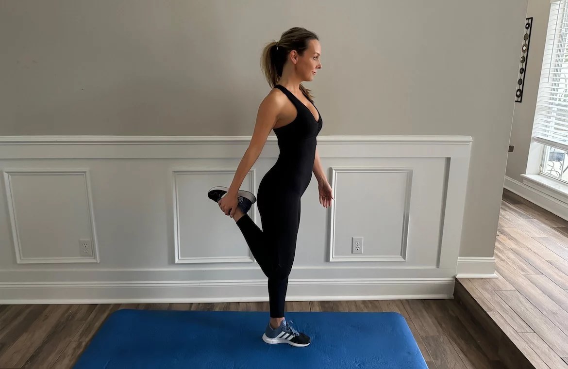 female PT with a long blond ponytail wearing black workout onesie shows the correct form for the standing quad stretch