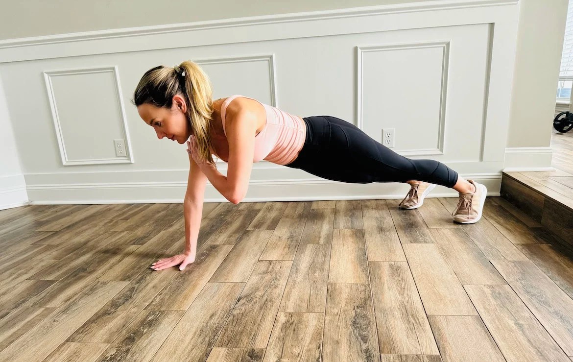 female PT with a blond ponytail wearing a tank top and leggings shows how to do shoulder tap push-ups with correct form