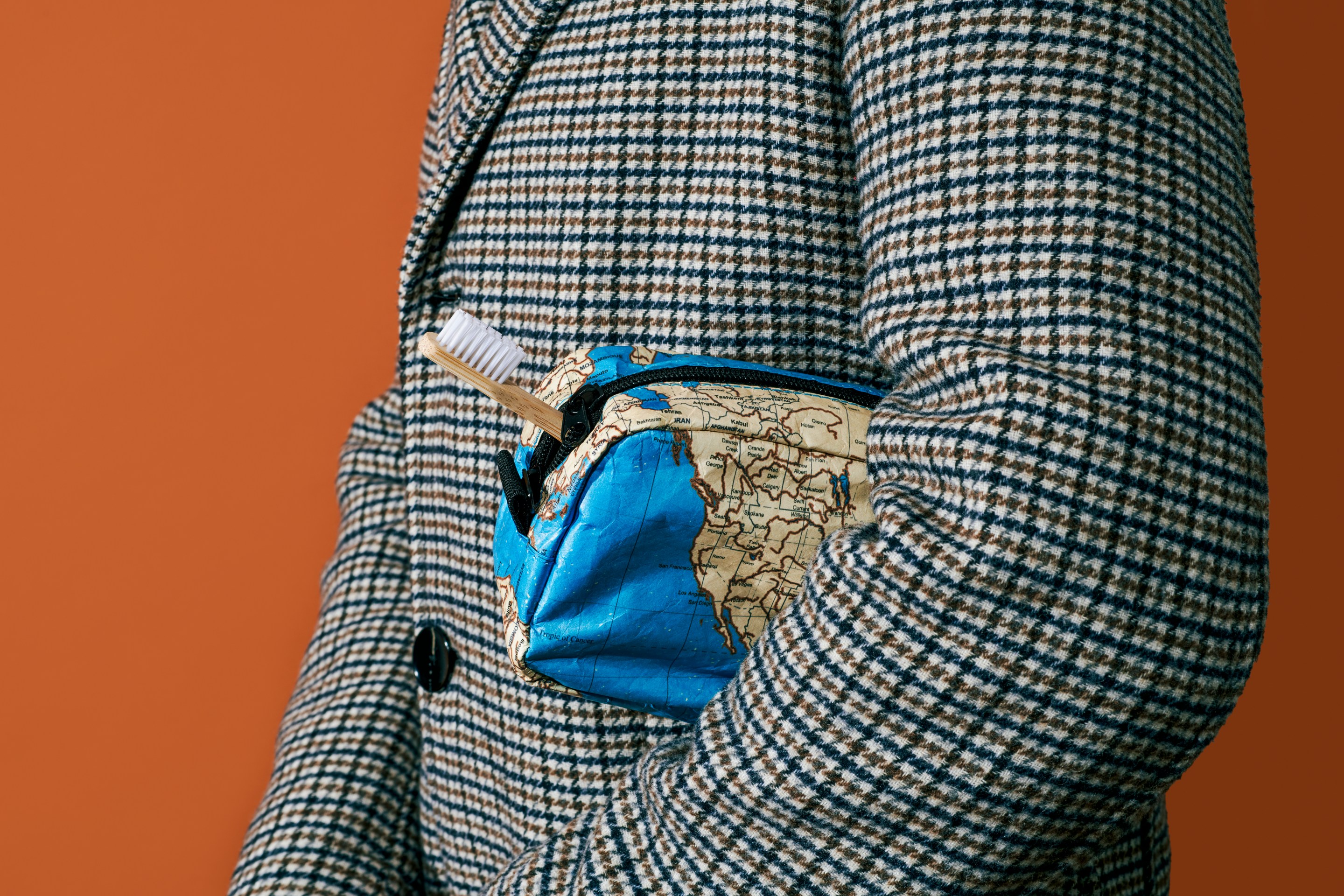 A person in a checkered blazer holding a world map-patterned toiletry bag with a toothbrush sticking out of it underneath their arm.