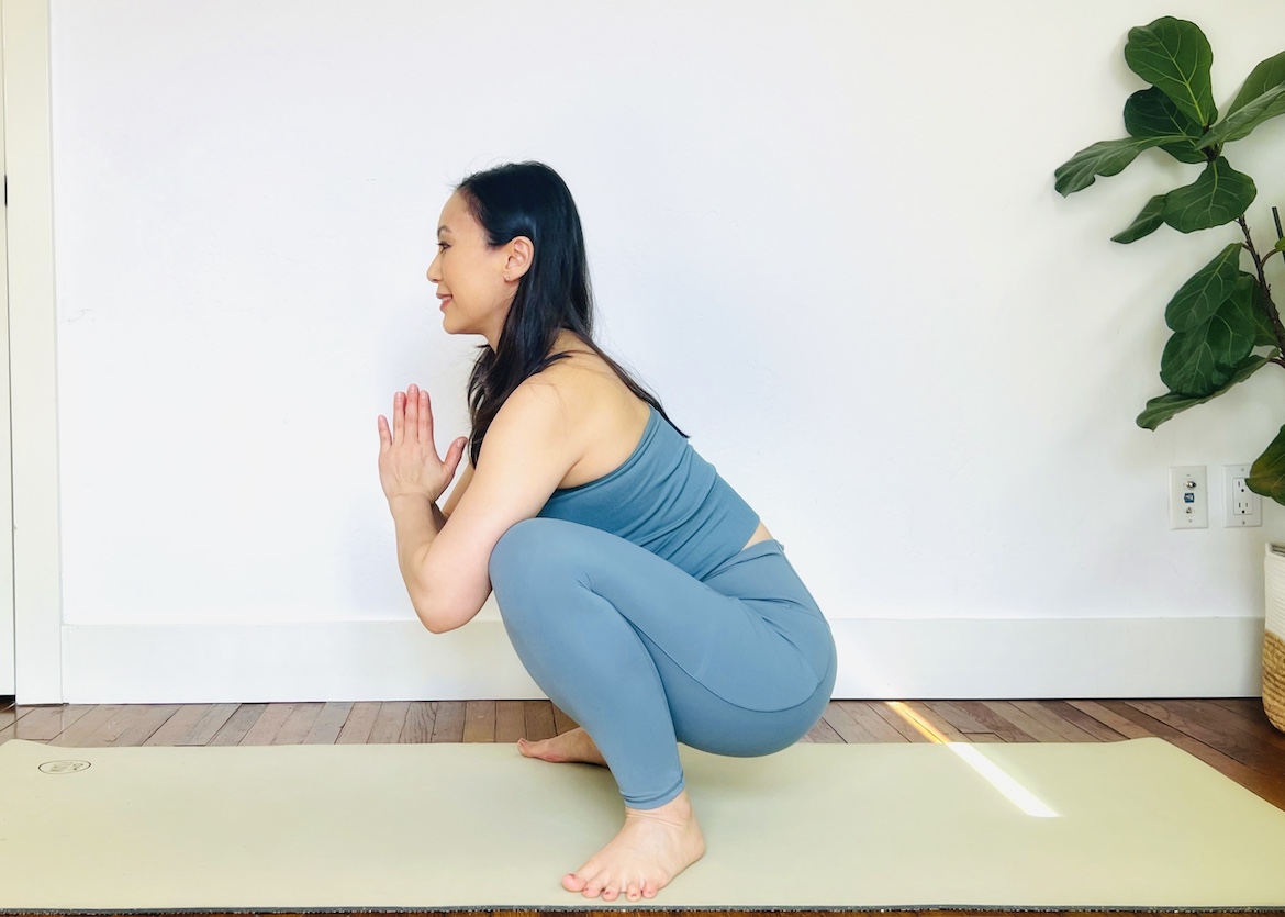 female yoga teacher wearing a matching blue set squats on her mat in preparation for crow pose