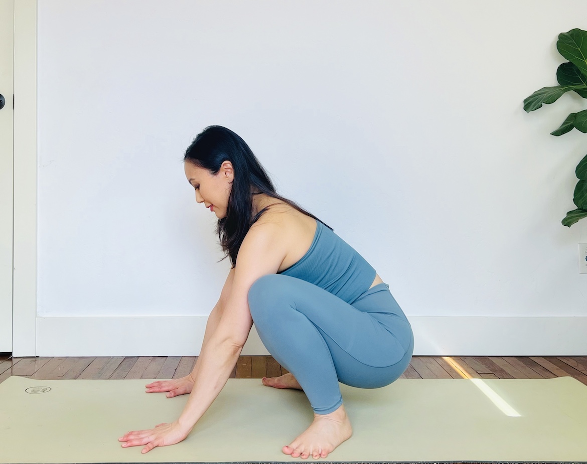 female yoga teacher wearing a matching blue set squats on her mat with her hands on the map and knees on her triceps in preparation for crow pose