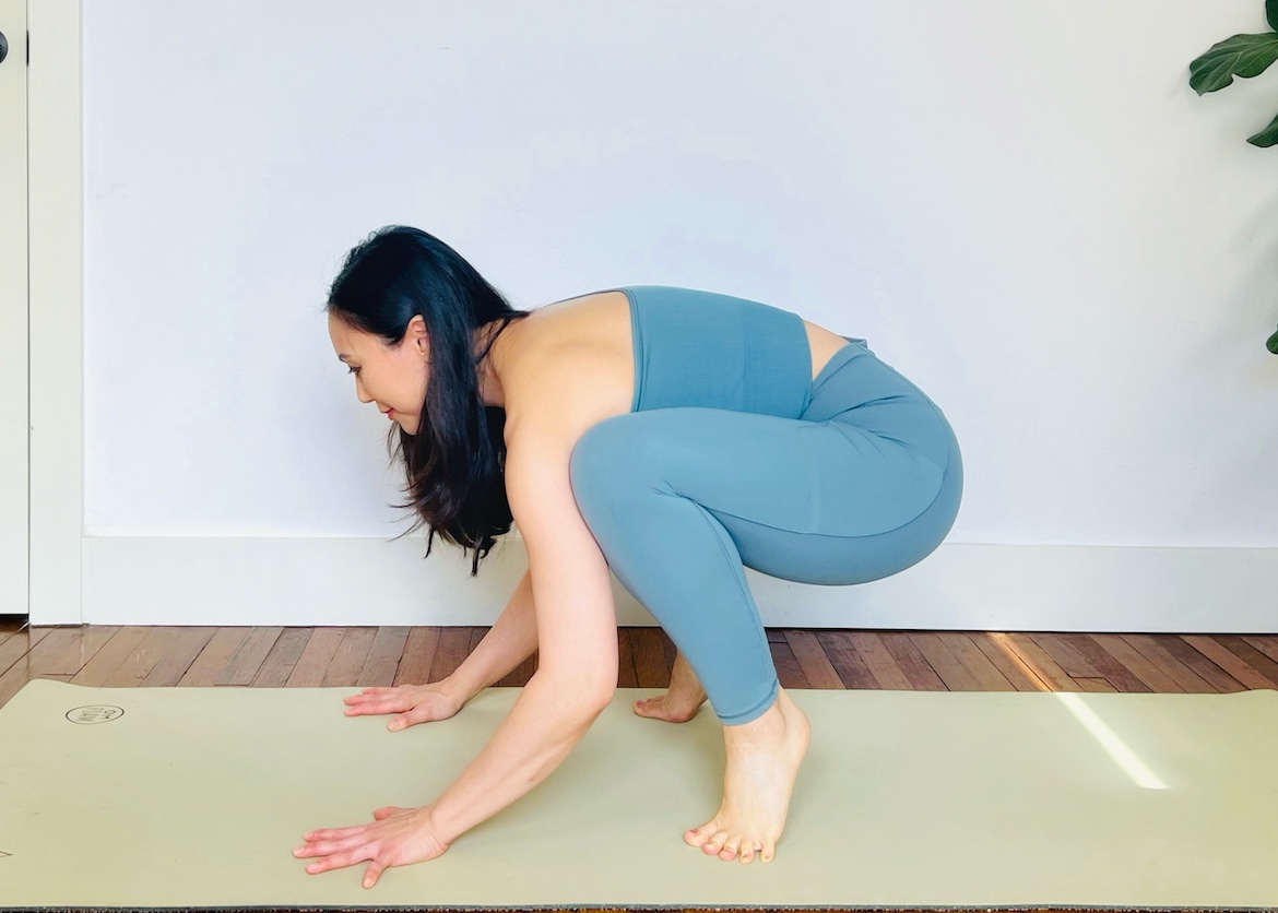 female yoga teacher wearing a matching blue set squats on her mat with her hands on the mat and knees bent, demonstrating an early step for crow pose