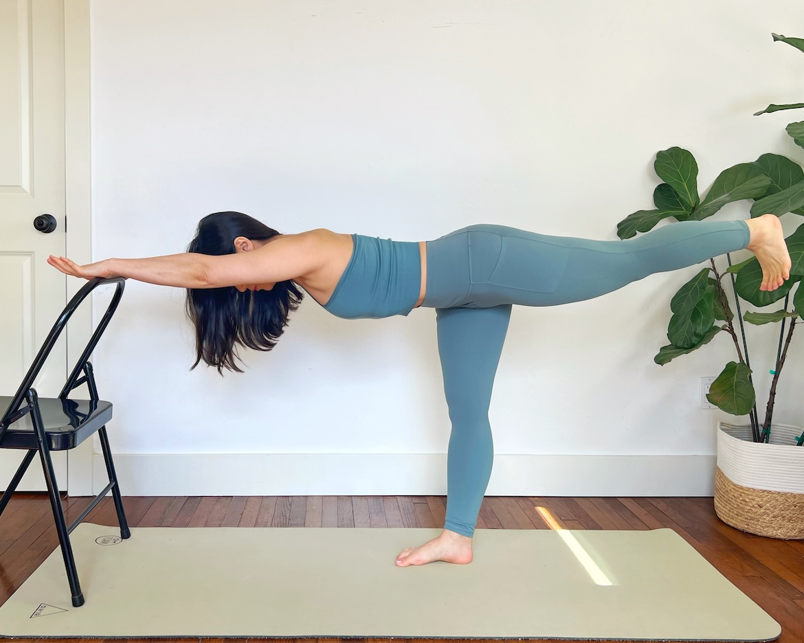 female yoga teacher with long dark hair shows how to modify warrior 3 with a chair in front 