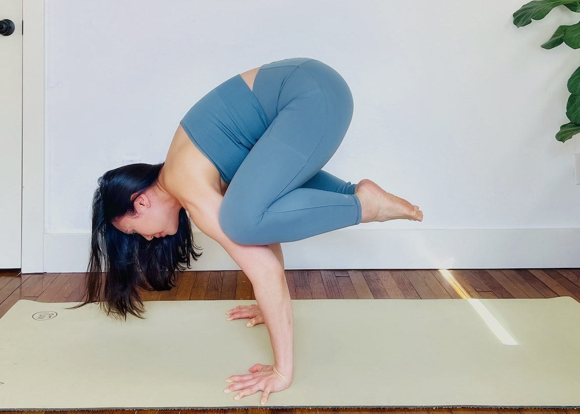 female yoga teacher wearing a matching blue set squats on her mat with her hands on the mat and knees bent, demonstrating an advanced crow pose form with straight arms