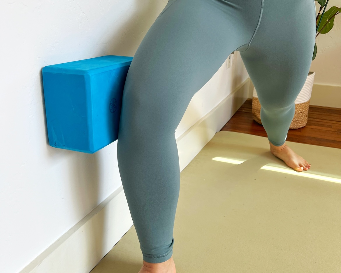 close up of a yoga instructor using a block in between the outside of her knee and a wall to help with warrior pose